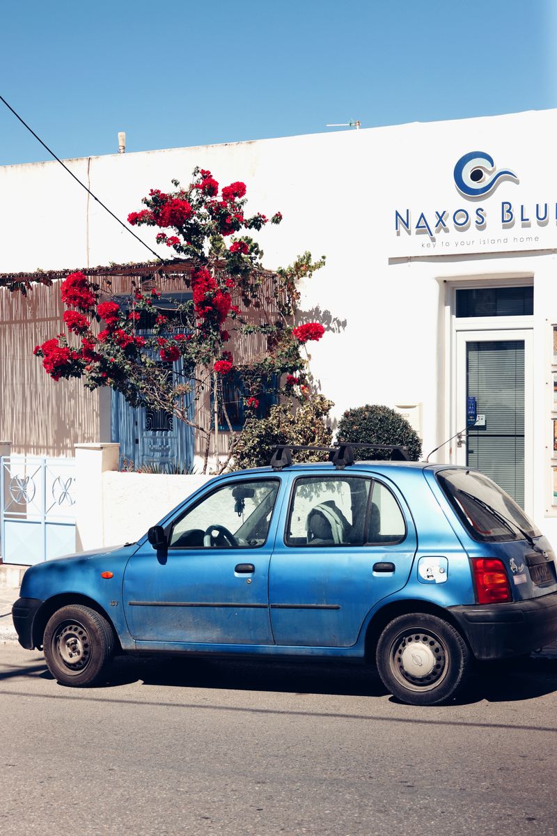 A picturesque scene in Naxos, Greece, featuring vibrant flowers and a blue car under a clear sky.