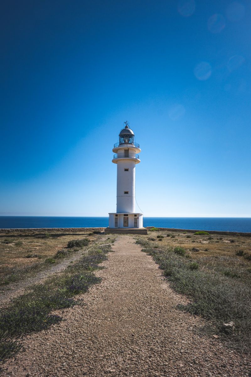 A lighthouse stands tall on a coastal path with a clear blue sky and sea in the background.